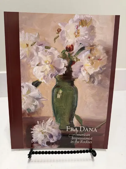 Fra Dana book resting on a bookstand with the text American Impressionist in the Rockies and a impressionist painting of a vase of white flowers on the cover