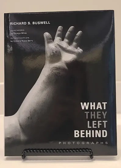 What they left behind book resting on a bookstand featuring a photograph of an aged doll arm missing a finger in black and white
