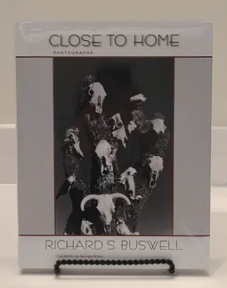 close to home book resting on a bookstand with the cover featuring a black and white image of cow skulls hanging on the trunk of a dead tree