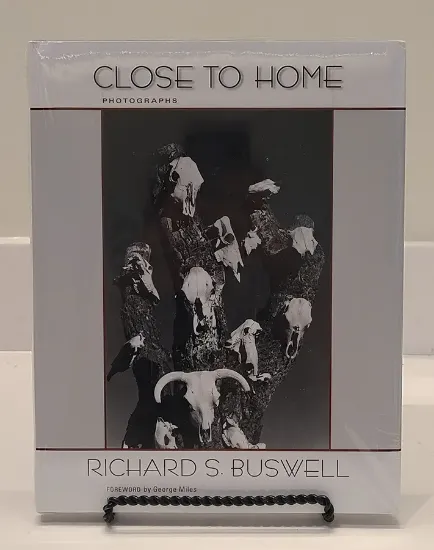 close to home book resting on a bookstand with the cover featuring a black and white image of cow skulls hanging on the trunk of a dead tree