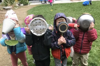 Four children outdoors hold pots and colanders in front of their faces, playing with kitchenware like masks.