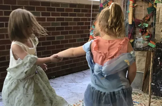Two children in dress-up outfits hold hands and spin near a brick wall, with colorful art materials hanging in the background.