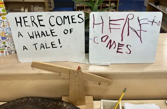 Two small whiteboards on a classroom table; the left reads “HERE COMES A WHALE OF A TALE!” and the right reads “HERE” with “COMES” scribbled underneath.