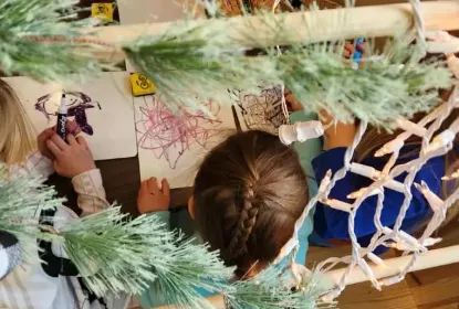 Overhead view of children drawing at a table, with green garland and string lights partially framing the scene.